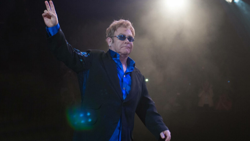 Elton John performing live in spotlight wearing dark suit bright blue shirt and signature glasses Wallpaper