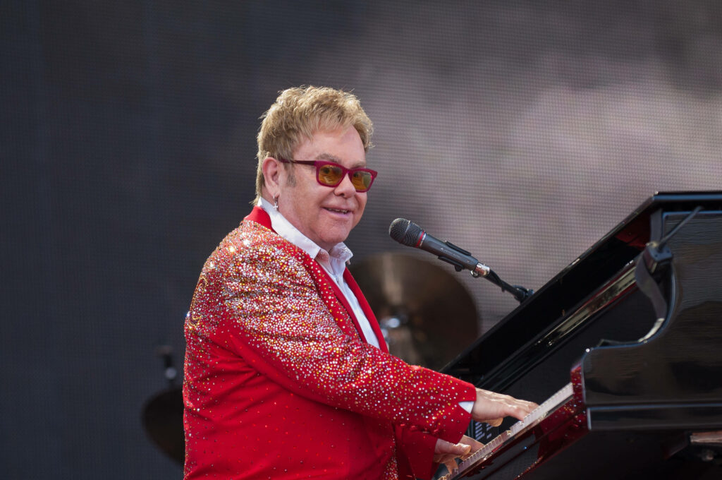 Elton John performing live at piano wearing red sparkling jacket and bold glasses vibrant wallpaper