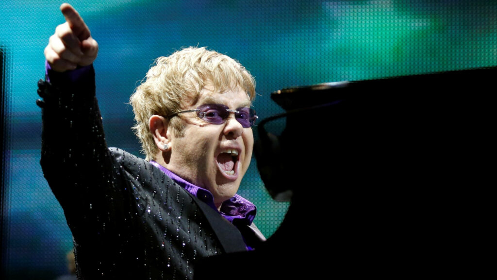Elton John performing live in sparkling attire and tinted glasses vibrant colorful stage background Wallpaper