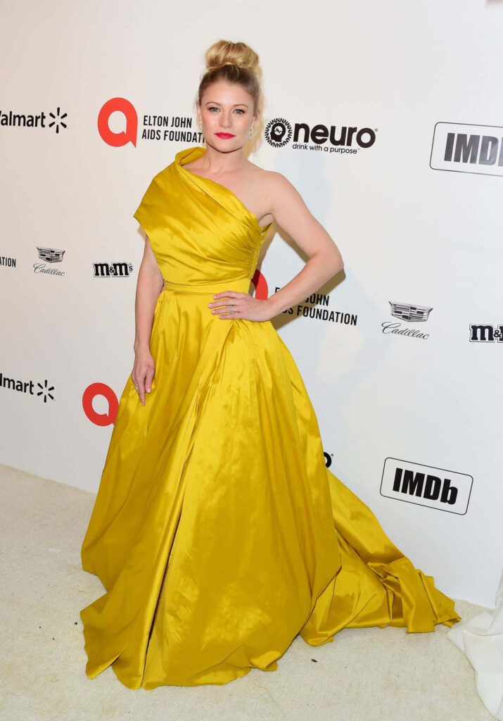 Emilie de Ravin in yellow off-shoulder gown posing on cream carpet with sponsor logo backdrop Wallpaper
