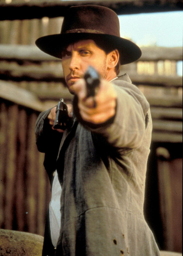 Emilio Estevez aiming revolver in Dollar For The Dead Western scene with rustic frontier background Wallpaper