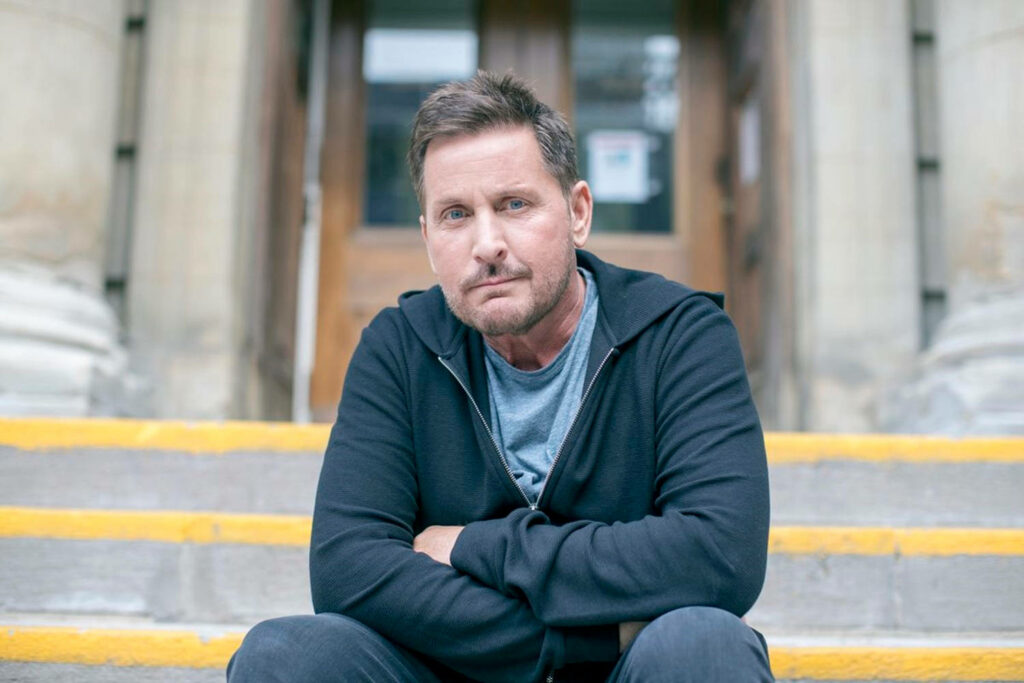 Emilio Estevez as Stuart Goodson sitting on stone steps outside public building in casual hoodie gray shirt Wallpaper