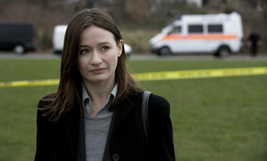 Emily Mortimer in professional attire with serious expression by police tape and emergency vehicles outdoors Wallpaper