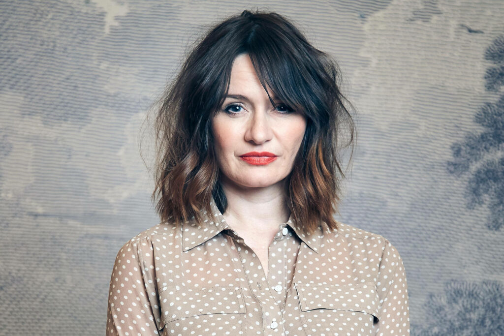 Emily Mortimer in sheer polka dot blouse with tousled hair and bold red lips against pastel backdrop Wallpaper