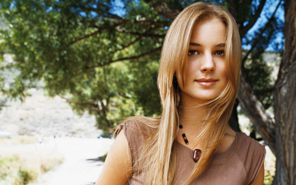 Emily VanCamp outdoors with lush green trees wearing brown top and beaded necklace wallpaper
