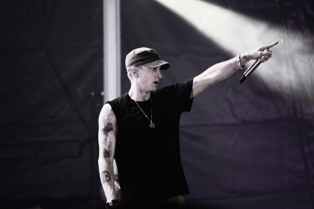 Eminem on stage wearing black t-shirt and cap with full sleeve tattoos holding microphone pointing crowd Wallpaper