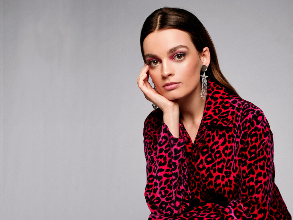 Emma Mackey in vibrant red pink leopard jacket against minimalist grey background with silver earrings Wallpaper