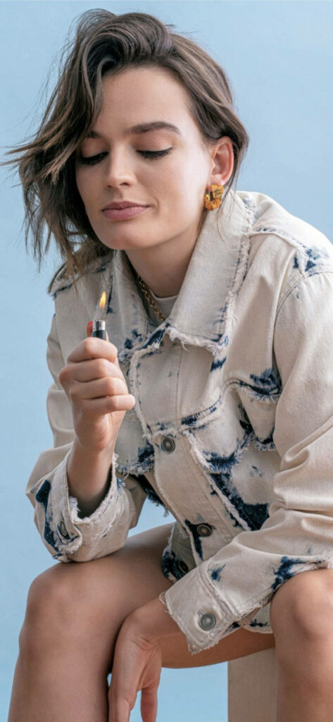 Emma Mackey holding lit lighter wearing trendy light-wash denim jacket with tie-dye accents Wallpaper