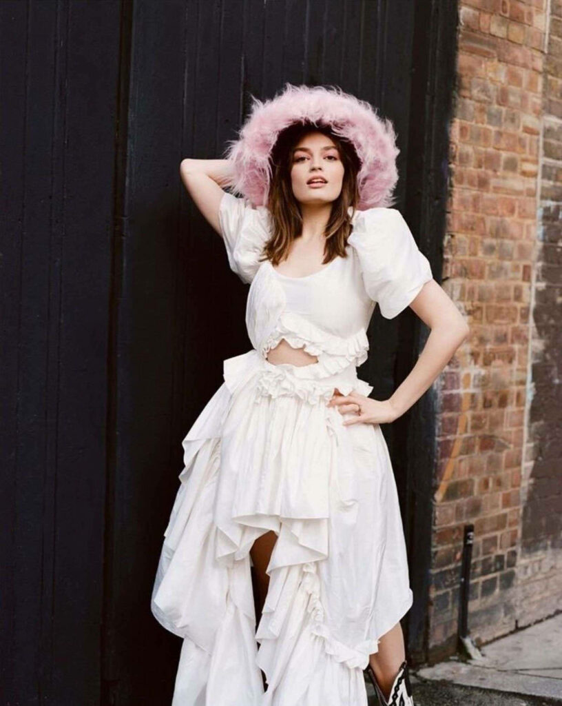 Emma Mackey in ruffled white dress and pink feathered hat urban brick wooden backdrop editorial Wallpaper
