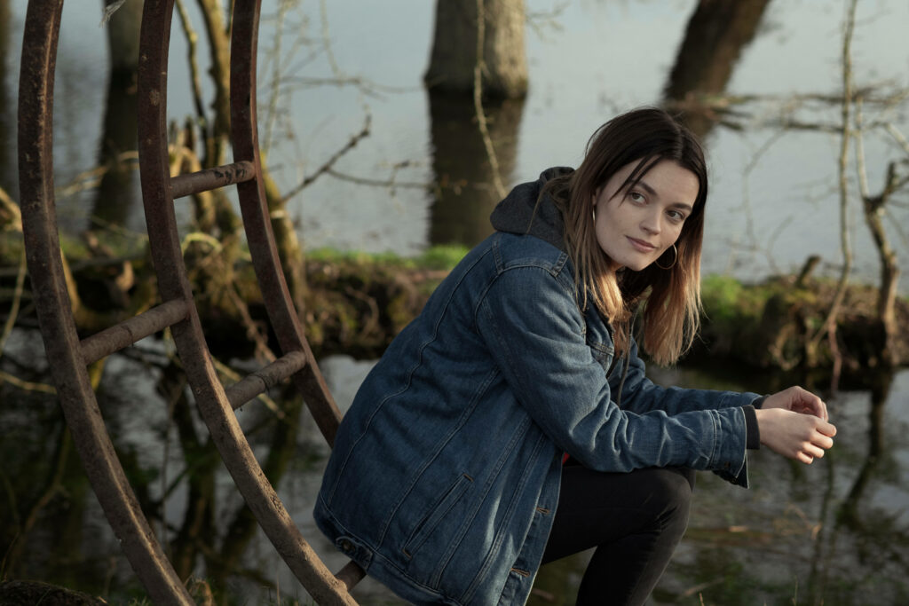 Emma Mackey in denim jacket reflects peacefully by calm lake with bare trees and soft natural light Wallpaper