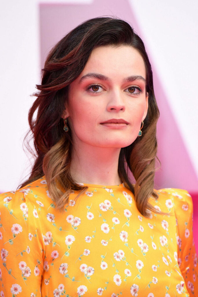 Emma Mackey in yellow floral dress with modern hairstyle against soft elegant wallpaper background