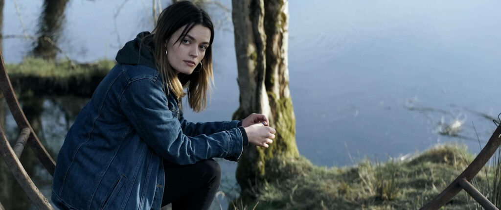 Emma Mackey as Maeve in denim jacket by calm water reflecting thoughtful mood wallpaper background