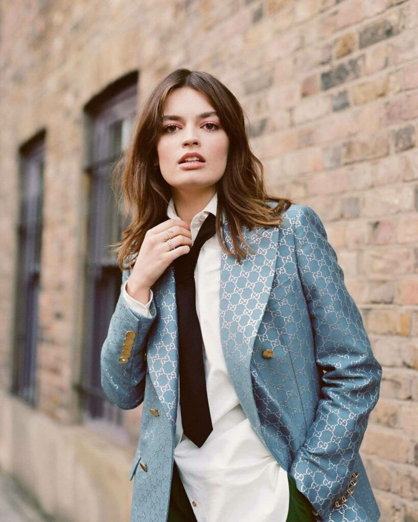 Emma Mackey in light blue Gucci monogram jacket white shirt black tie urban brick wall background Wallpaper