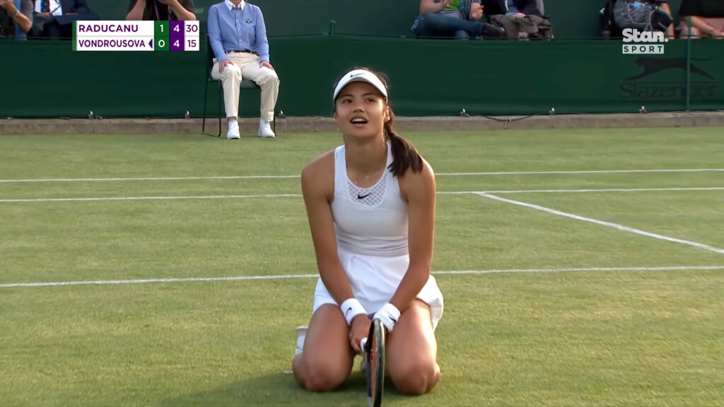 Emma Raducanu kneeling on Wimbledon grass court wearing white tennis outfit focused during match Wallpaper