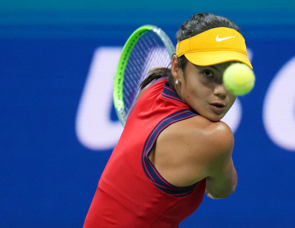 Emma Raducanu focused mid-motion returning tennis ball wearing red and yellow outfit on blue court Wallpaper