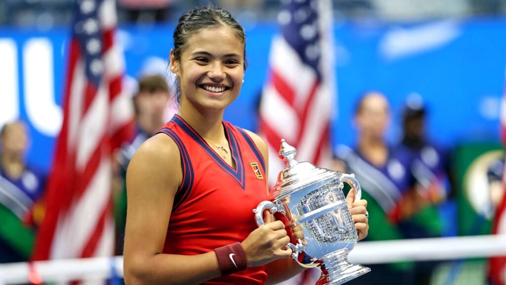 Emma Raducanu in red holding silver tennis trophy smiling courtside with American flags background Wallpaper