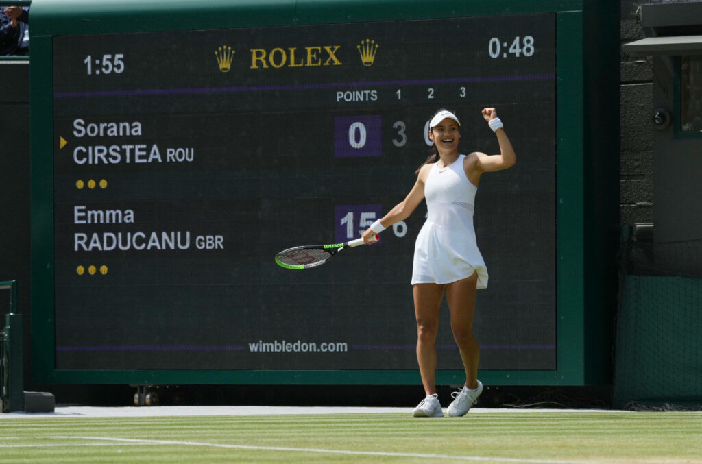 Emma Raducanu celebrates Wimbledon win in white tennis outfit on vibrant green grass court scoreboard visible Wallpaper