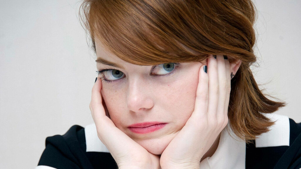 Emma Stone with bored expression resting face in hands blue eyes classic makeup neutral background Wallpaper