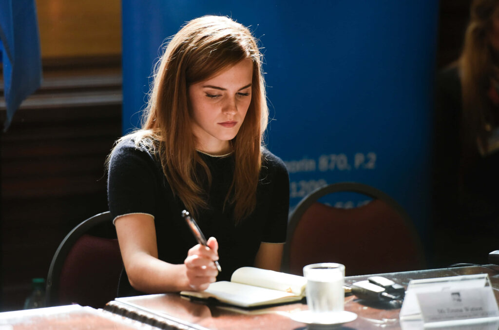 Emma Watson writing thoughtfully in notebook at table with soft natural light and blurred background Wallpaper