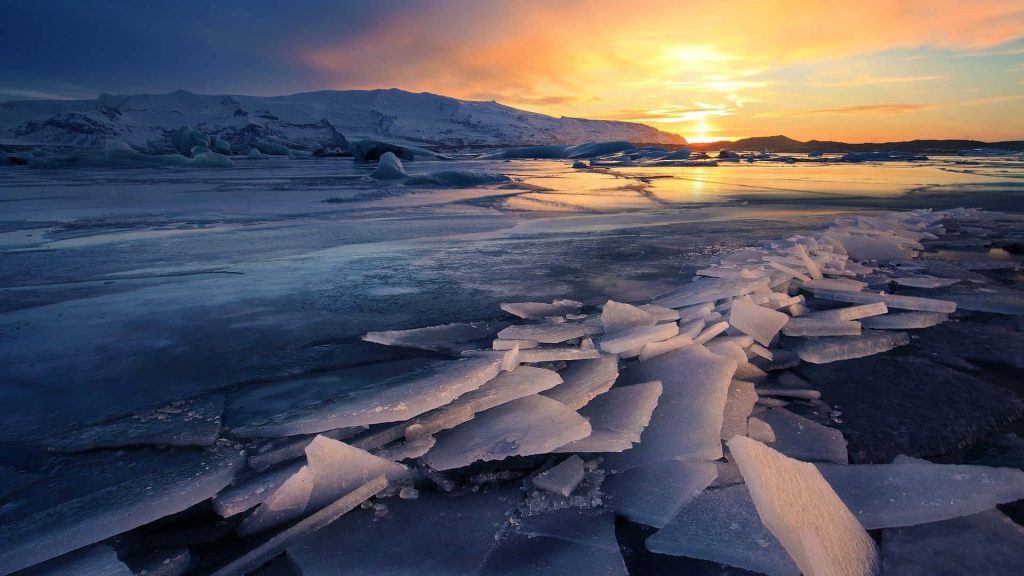 Enchanting winter sunset over frozen lake with jagged ice shards and snowy mountains background Wallpaper