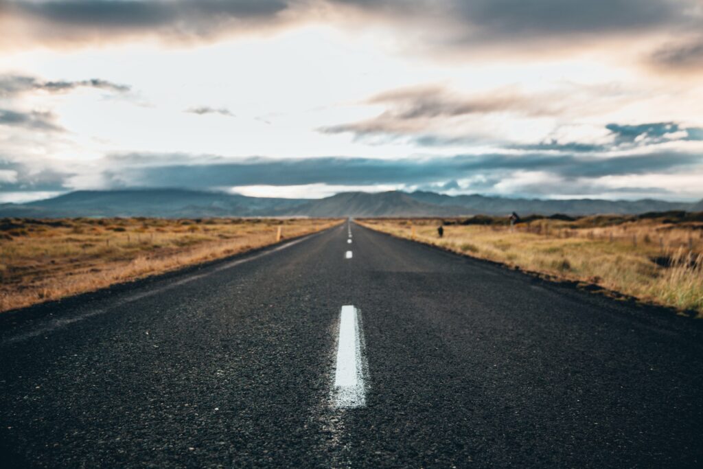Straight asphalt road through golden grasslands and mountains under dramatic cloudy sky wallpaper