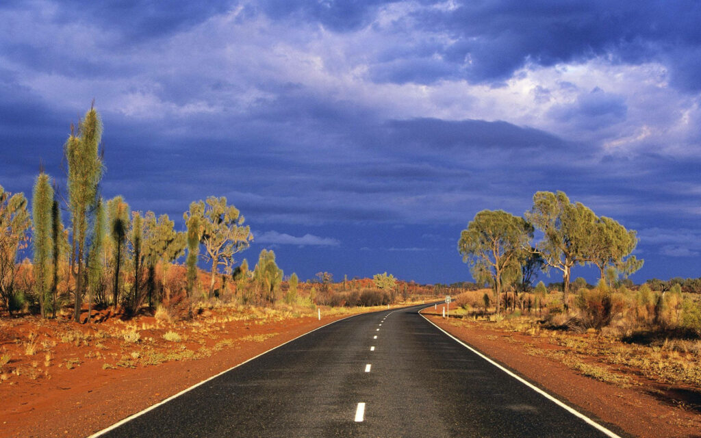 Long asphalt road winding through rugged Australian Outback with red earth and blue skies wallpaper