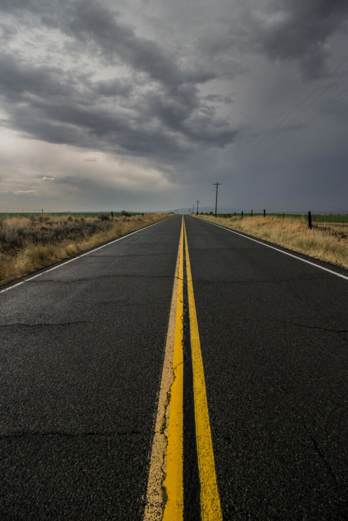 Endless asphalt road with yellow markings under cloudy sky bordered by dry grass wallpaper background