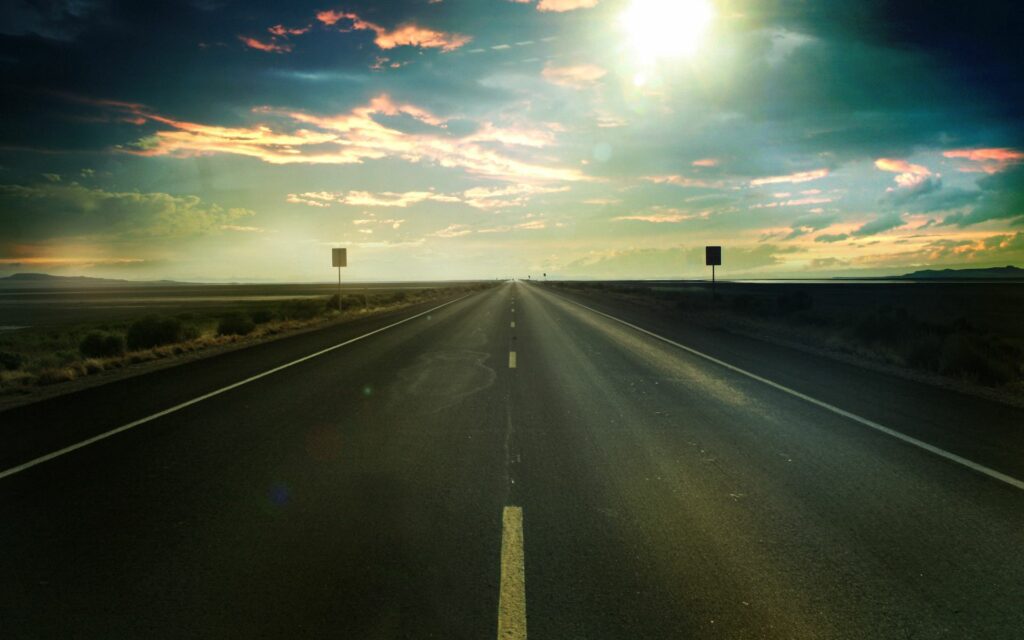 Asphalt road with white markings stretching to horizon under dramatic sky and soft sunlight Wallpaper