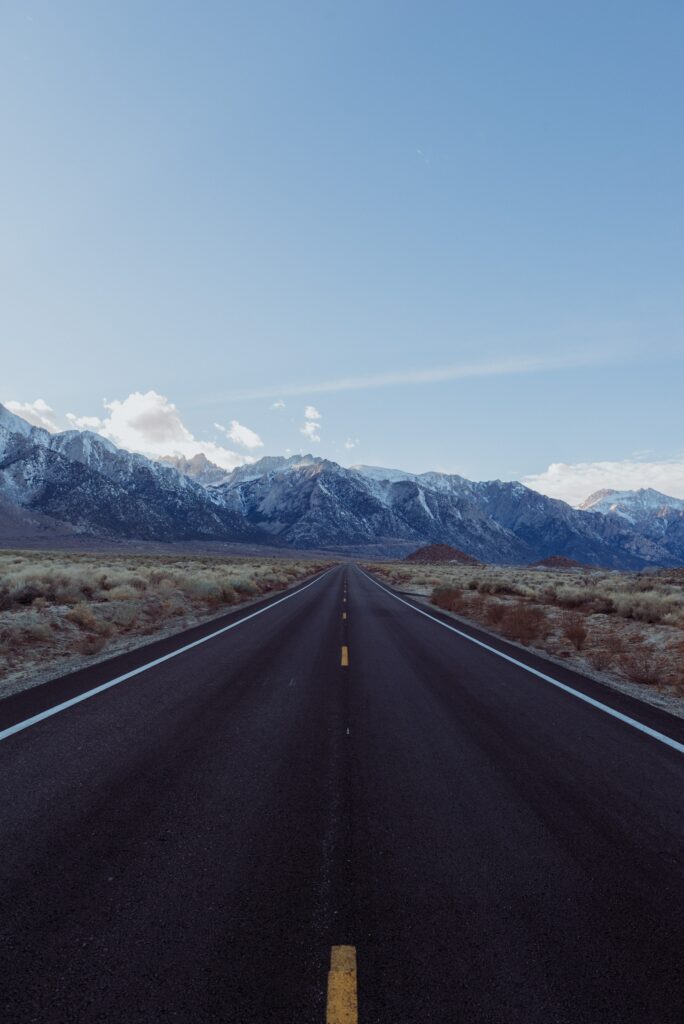 Straight asphalt road through dry grasslands towards snow-capped mountains under clear sky wallpaper
