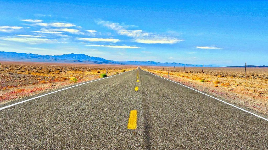 Endless asphalt road through vast desert toward misty mountains under bright daylight sky Wallpaper