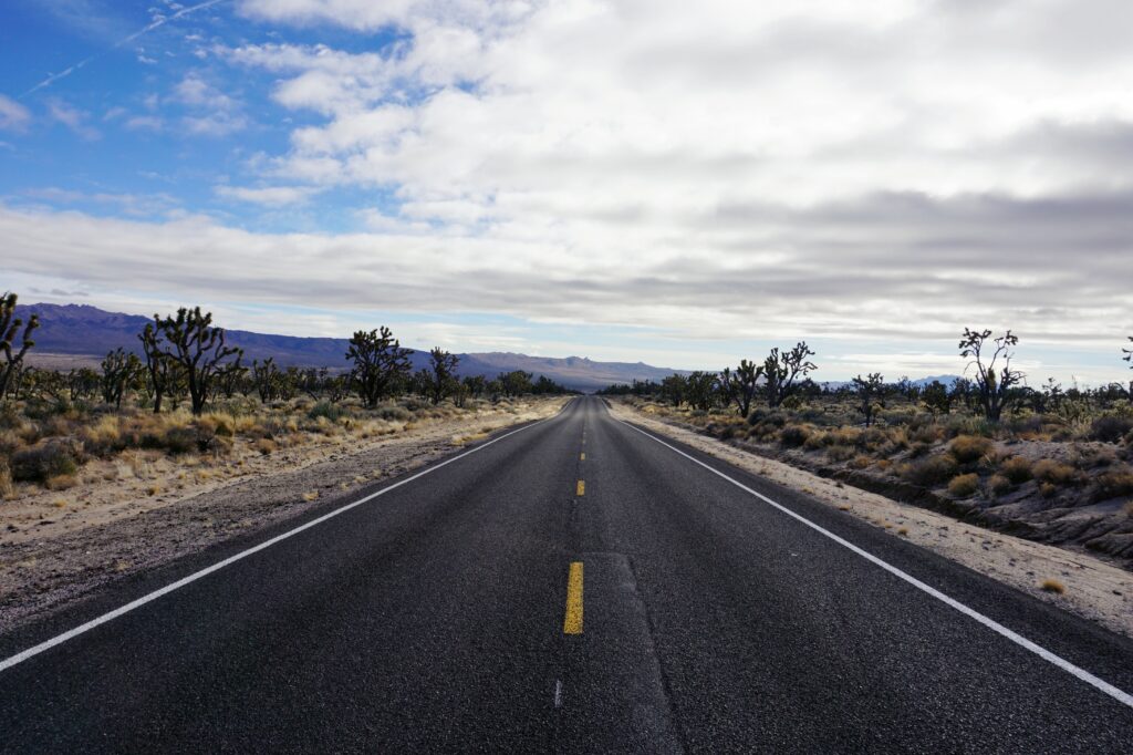 Long asphalt road with yellow lines through desert and Joshua trees under partly cloudy sky wallpaper