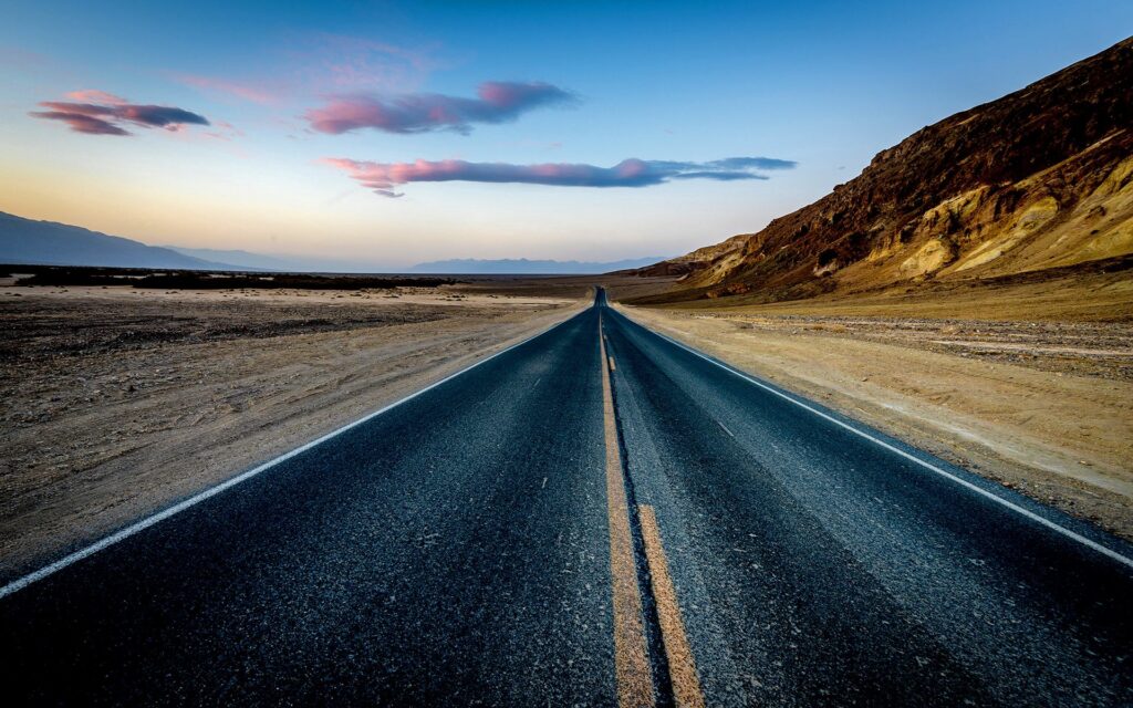 Long empty desert road with sandy dunes rugged hills and pink clouds under vibrant sky wallpaper