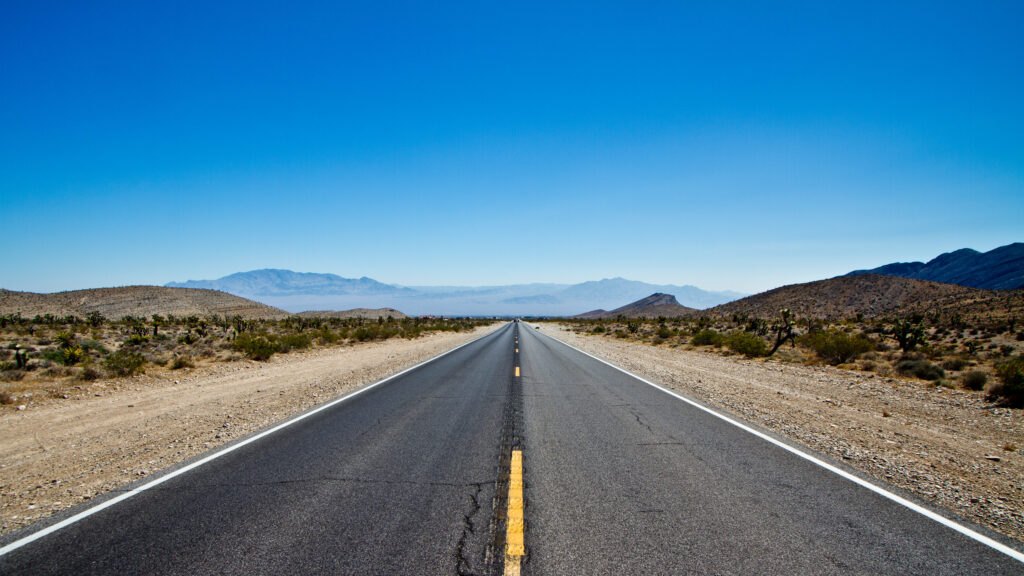 Long straight desert road with yellow center line stretching to distant mountains under blue sky Wallpaper