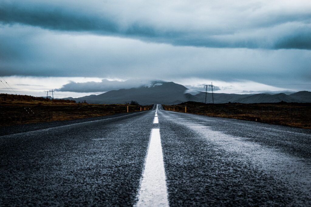 Long asphalt road with white markings leads to mountain under moody cloudy sky rugged terrain background Wallpaper