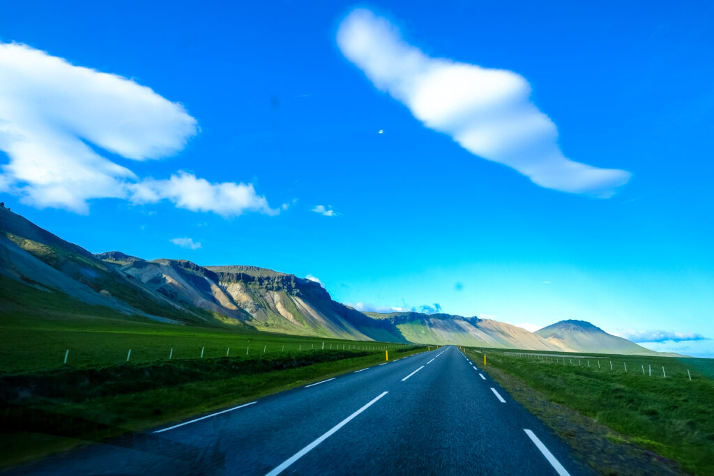 Open road through green fields and mountains under blue sky with clouds scenic adventure view Wallpaper