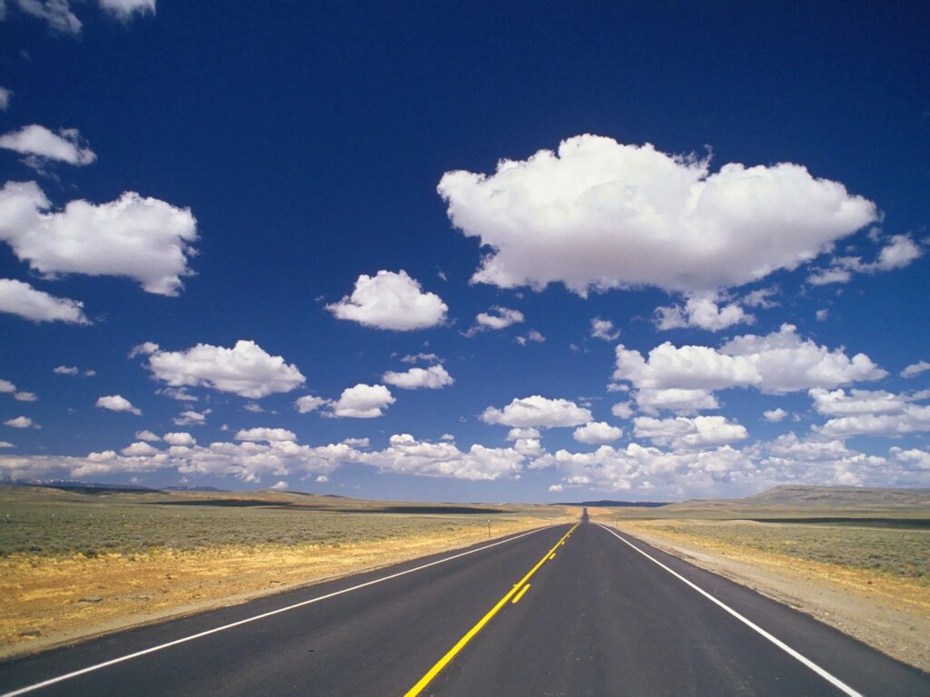 Straight road with yellow lines through dry landscape under blue sky with white clouds wallpaper