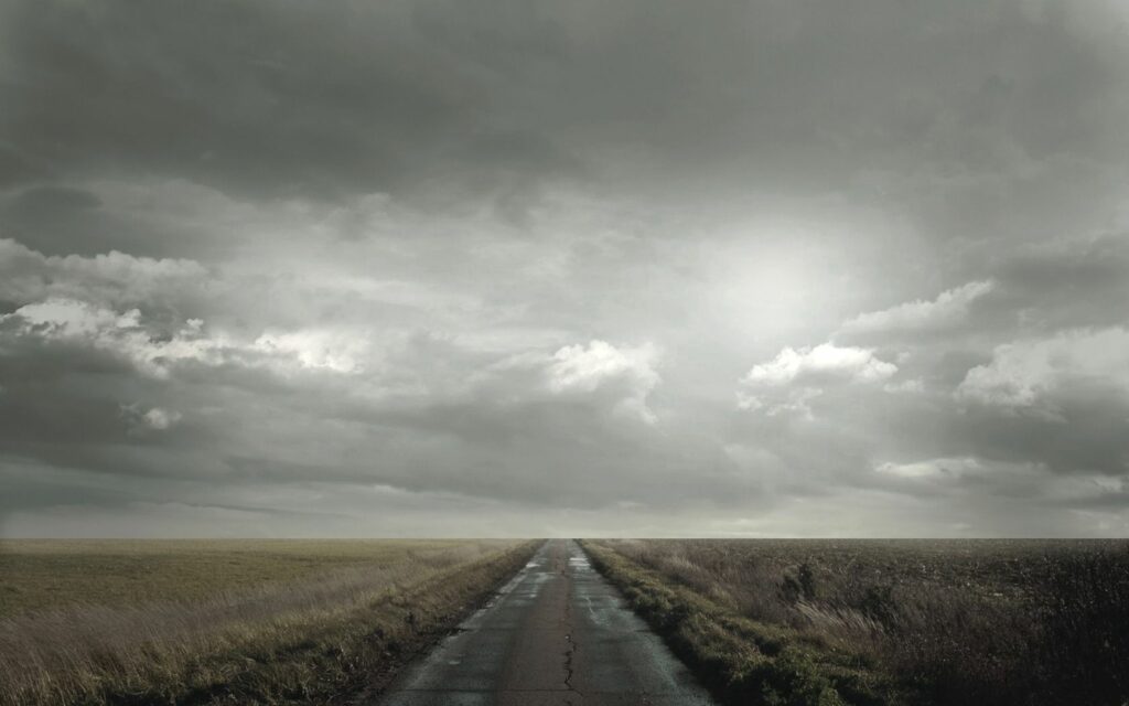 Straight road through open fields under dramatic cloudy sky moody landscape wallpaper background