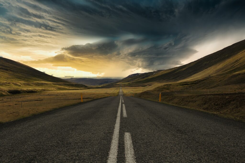 Asphalt road through rolling hills under dramatic cloudy sky with golden sunlight at horizon Wallpaper