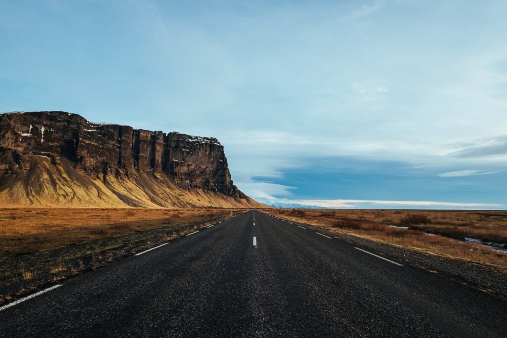 Long straight road with white markings beside golden grasslands and hills under a wide sky Wallpaper