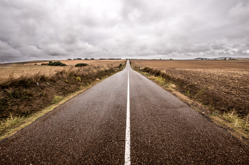 Endless straight road through open fields under dramatic cloudy sky showcasing solitude and adventure Wallpaper