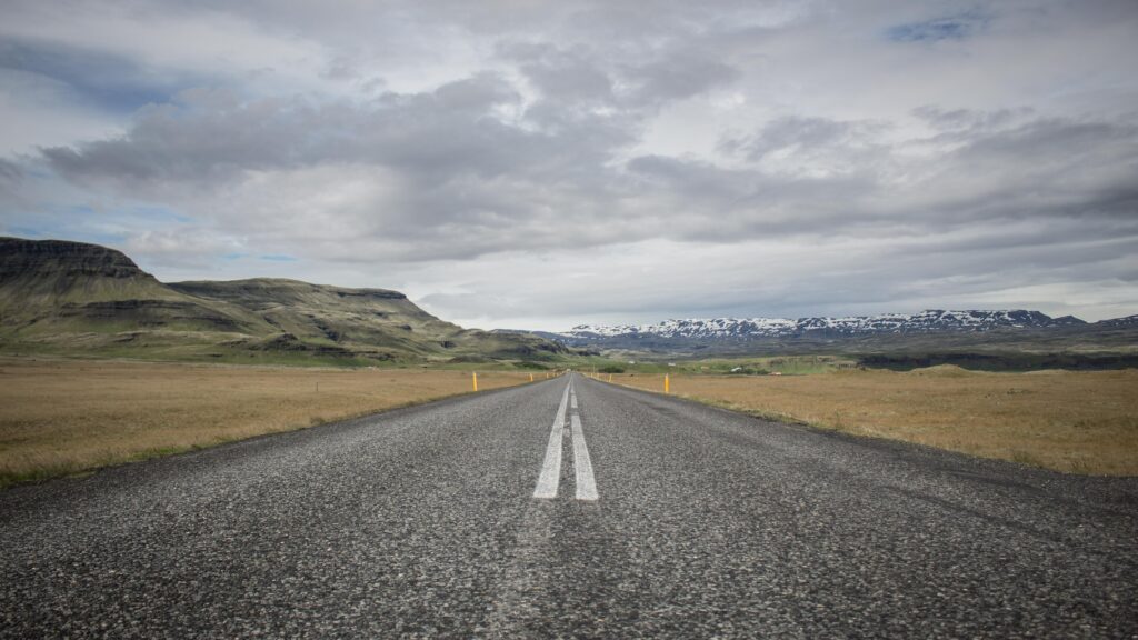 Long straight asphalt road with white markings through fields and hills under cloudy sky wallpaper