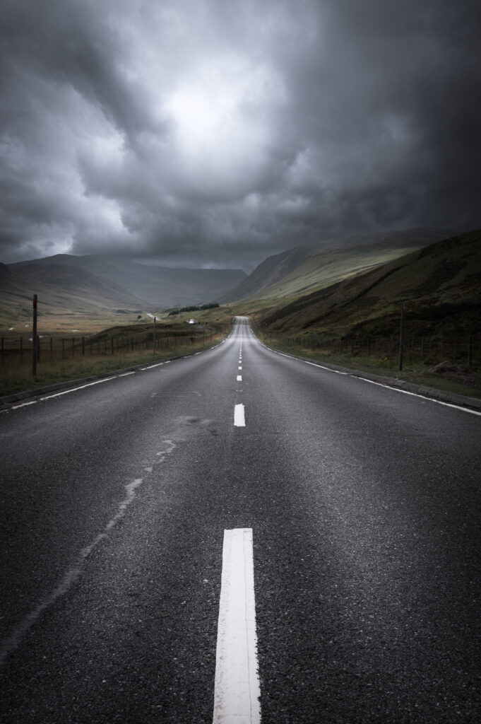 Long straight road with white markings under dramatic overcast sky leading to misty hills Wallpaper
