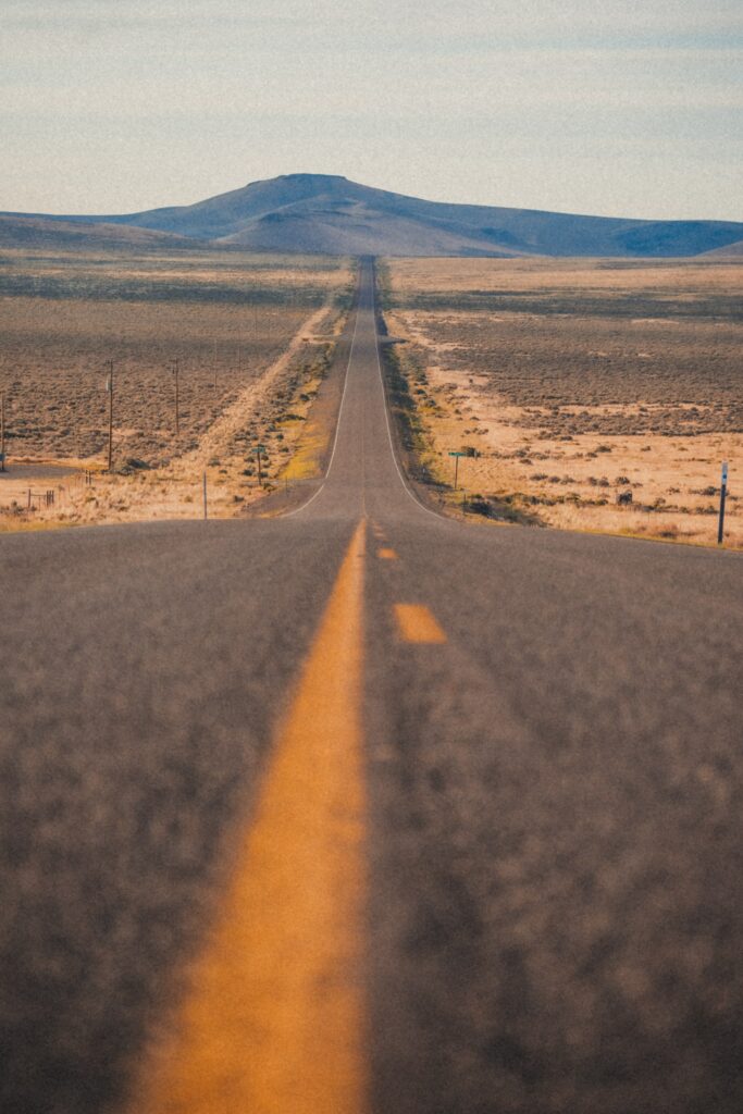 Long straight road with yellow centerline leading to mountains under pale blue sky wallpaper background