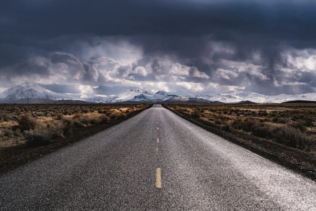 Long straight road toward snow-capped mountains under dramatic cloudy sky with rugged terrain Wallpaper