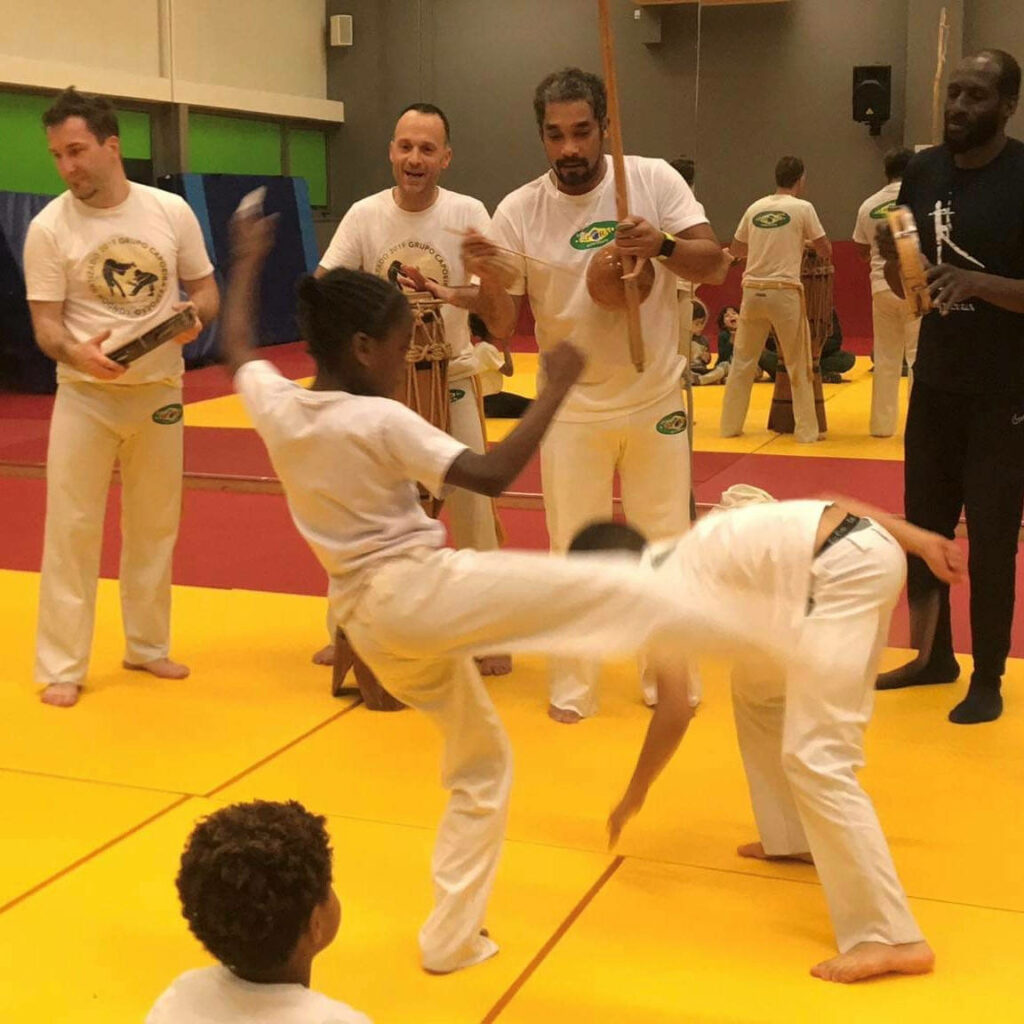 Children practicing energetic capoeira moves on bright mat with instructors playing traditional instruments Wallpaper