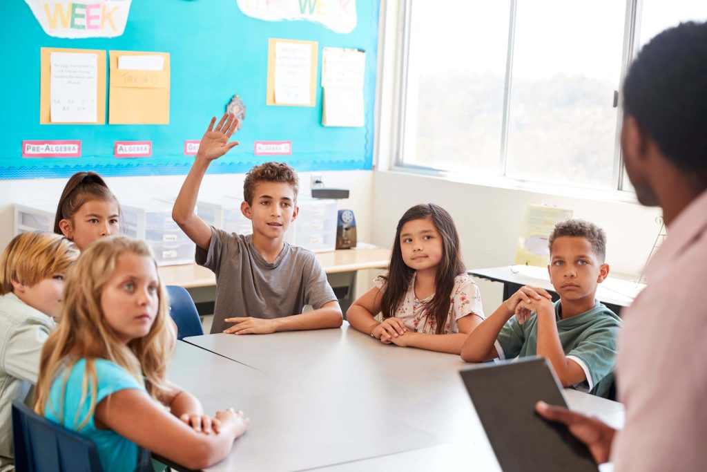 Diverse elementary students learning attentively in bright classroom with teacher and raised hand Wallpaper