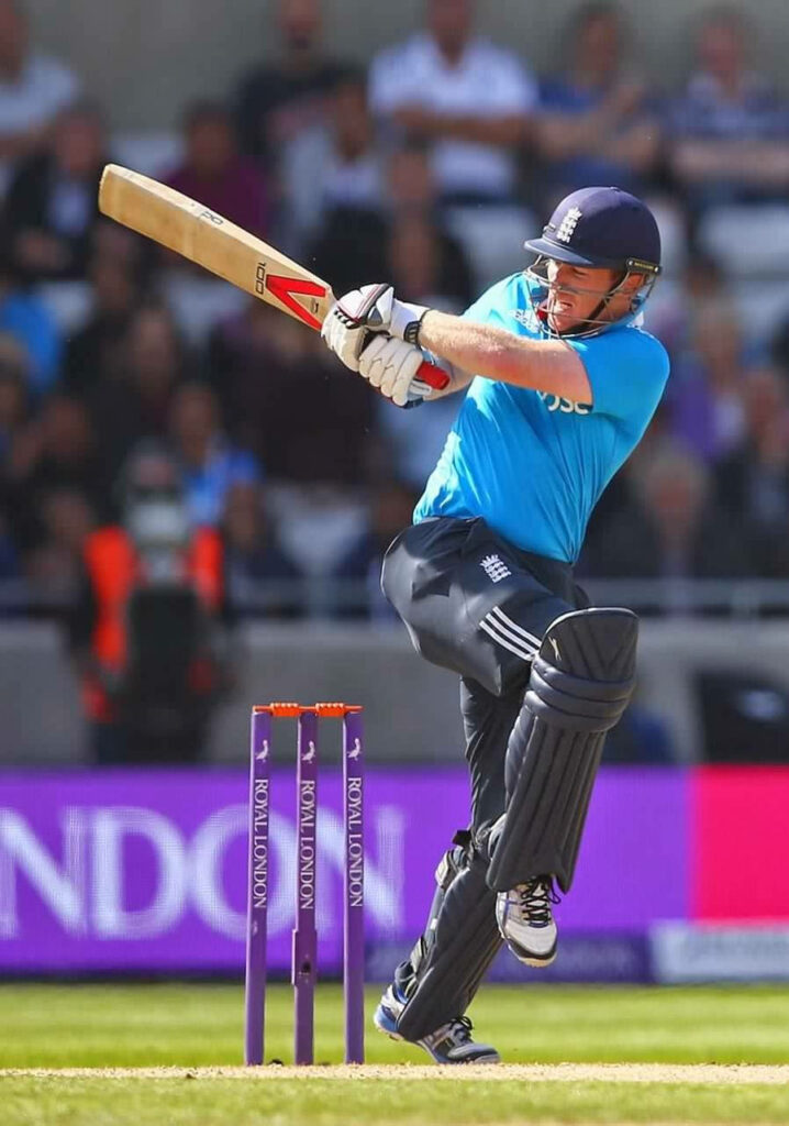 Eoin Morgan batting in England blue kit with raised knee stadium background dynamic cricket action Wallpaper