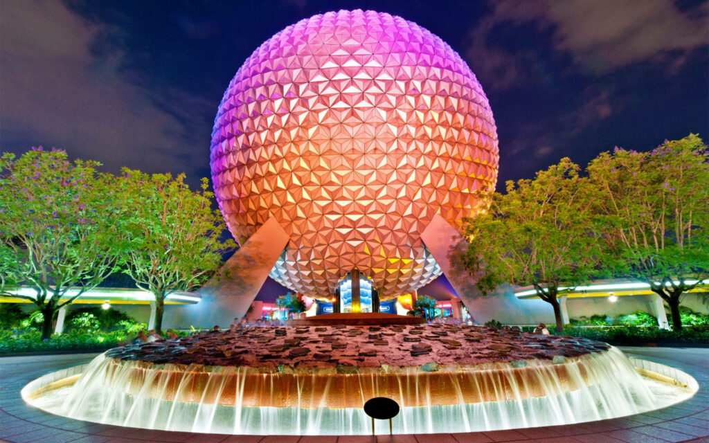 Epcot sphere glowing orange and purple at night with lush greenery and cascading fountain background Wallpaper