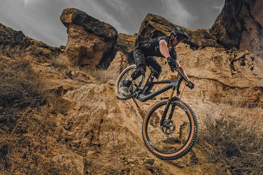 Cyclist on black brown mountain bike rides rugged rocky trail with dramatic cliffs and moody sky Wallpaper