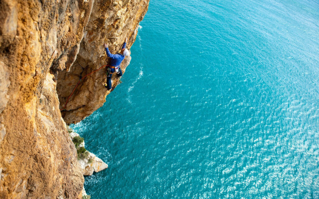 Climber ascending rugged cliff with panoramic sparkling blue sea adventure nature wallpaper
